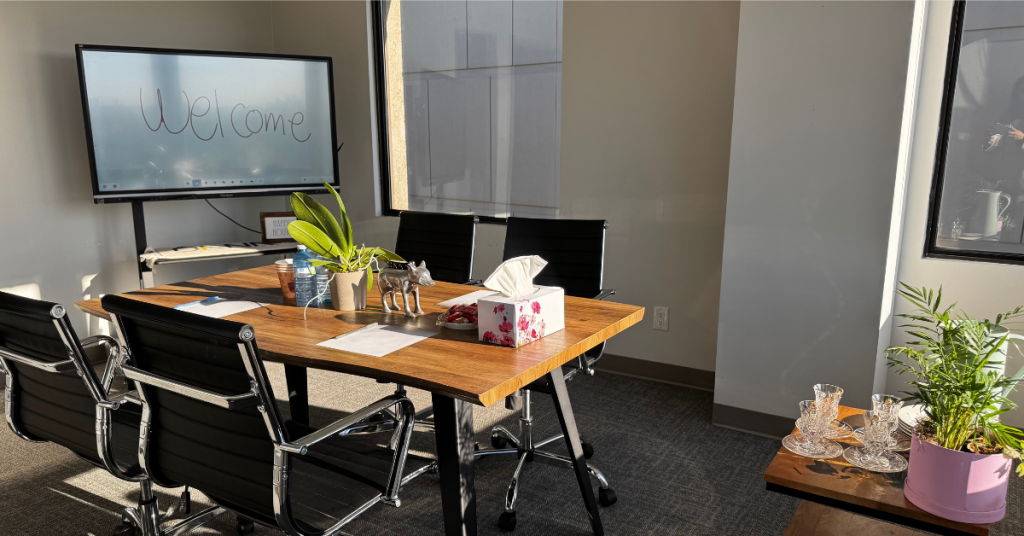 A sunlit meeting room at Luminic Studio with a wooden table, black chairs, a large display screen and plants, available for shared office members in the GTA